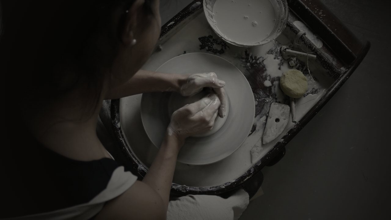 Seen from above, a potter centres clay on the wheel beside a bucket of slip, a sponge and a wooden rib