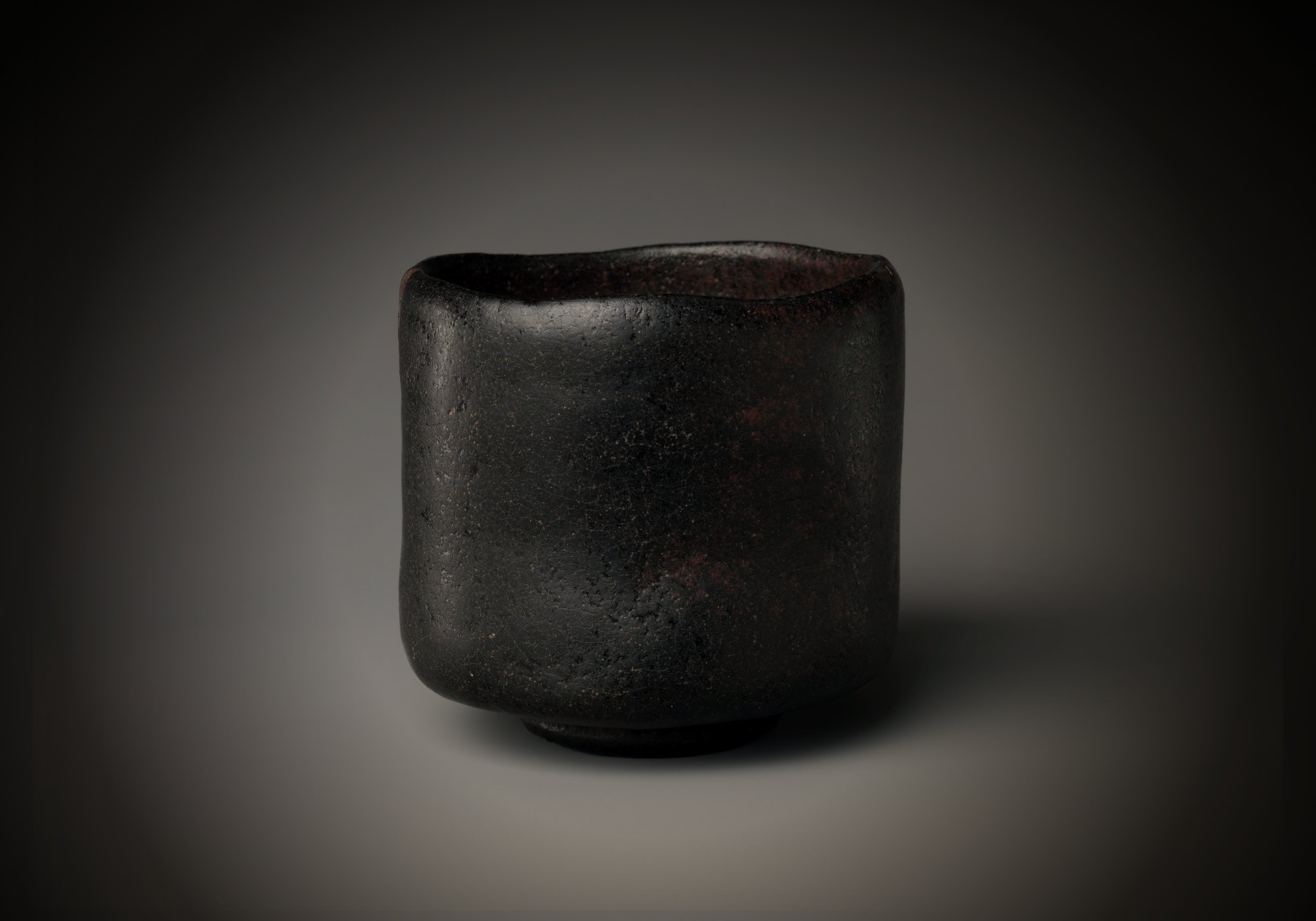 A black tea bowl with a soft, uneven rim, lit from the left; a line of gold traces a crack down its side